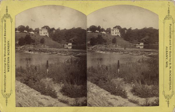 Stereograph of a spillway on a river. Above, on the hill, is a home and tents pitched behind it. An American flag is flying. Below is an excursion boat, painted to look like a sidewheel steamboat, on the pond created by the spillway. In the foreground is a stone wall, stone structure, fence post and river below the spillway. Printed on the card: "Wandering Among the Wonders and Beauties of WESTERN SCENERY."