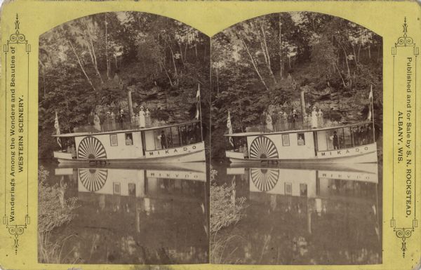Stereograph of the "Mikado," an excursion boat painted to look like a paddle wheel steamboat. It is stopped along the shoreline of the river where rock formations and trees are covering the bank. Most of the passengers are on the boat, but several have climbed the bank. The group of people are facing the camera. There are flags on the bow and stern.