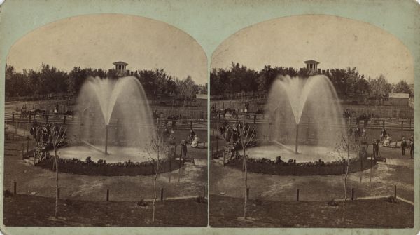 Stereograph of Artesian well. A stone wall surrounds the well, with an observation area beyond. Groups of visitors are milling about and a drive with horses and buggies is behind the well. In the background are buildings and trees. Information from the reverse: "Artesian Well. cor. Bluff & Wis. St. Flow of water, 30,000 bbls. per day. Height of pipe 10 ft. Was begun in Nov. '75 and finished in Feb. '76. Its depth is 960 ft. through 147 ft. of sand to solid rock about 2 ft. of hard limestone, then 107 ft. of soft slate or slale of a greenish color, then a soft white sand rock in which at a depth of 272 ft. the first flow of water was obtained. The sand rock continued with an occasional strata of slate & thin layers of a harder limestone until at about ?4? Ft. a sort of conglomerate sand rock was reached which yielded the most water. It blongs (<i>sic</i>) to the saline order of mineral waters and is very beneficial in diseases of the kidneys, bowels and all chronic diseases. Address Artsian Well Co. for pamphlet."