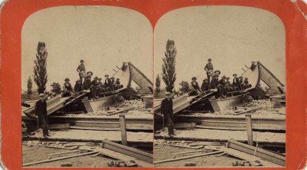Stereograph of the wreckage of a building in the aftermath of a cyclone. A group of men and boys are posing standing or sitting for the photograph. In the foreground is the platform of what was the porch. Trees and intact buildings are in the background. On the reverse: "Views of the Cyclone at Oshkosh, Wisconsin, July 8th, 1885."