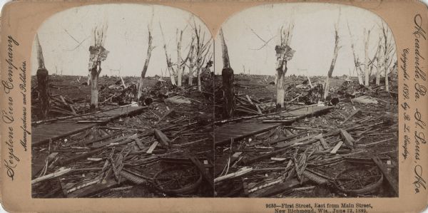 Stereograph of the devastation caused by a tornado. All that remains of trees are the main branches and trunks, some with building debris caught in them. Pieces of agricultural implements are in the foreground. Piles of branches, boards and other building materials cover the ground. Behind the tree trunks on the right is an upside down wagon. In the background are partially intact structures. Caption at foot: "9653 — First Street, East from Main Street, New Richmond, Wis., June 12, 1899."