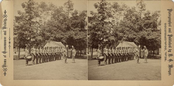 Stereograph of a line of soldiers standing at attention in formal uniform. Most of the soldiers are carrying weapons with bayonets. The soldier on the left has a drum. In front of the line, one soldier is saluting another. Trees and a large tent are in the background. Text printed on front: "Views of Oconomowoc and Vicinity. Photographed and Published by D.G. Munger, Oconomowoc, Wis."