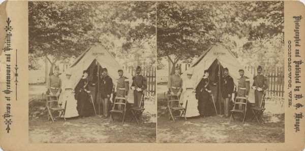 Stereograph of four soldiers and two women posing in front of a tent, with a sign above the door that reads: "Adjutant." One woman is wearing a sash and is holding a sword with the point on the ground. Two soldiers have their hands resting on folding chairs and a small table is sitting to the right. In the background are a fence and more tents with trees overhead. Text printed on front: "Views of Oconomowoc and Vicinity. Photographed and Published by D.G. Munger, Oconomowoc, Wis."
