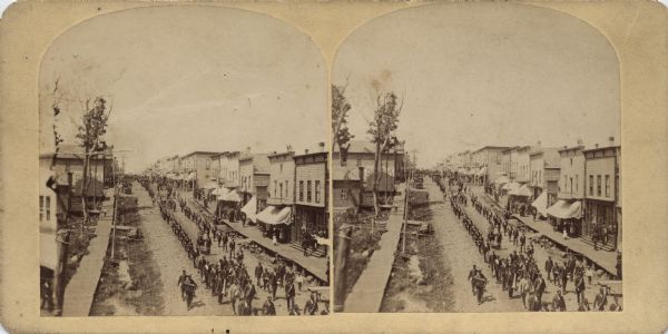 Stereograph of an elevated view of a parade marching through town. Many of the participants are wearing uniforms, and some are carrying musical instruments. The street is unpaved, and there are boardwalks on both sides. Many of the buildings have awnings. Hand-written on reverse: "Parade in Hurley."