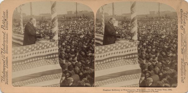 Stereograph of President McKinley speaking to a large crowd from a stage decorated with bunting. In the background are buildings and train cars. Caption reads, "President McKinley at West Superior, Wisconsin, — On the Western Tour, 1899. Copyright 1900 by Strohmeyer & Wyman."