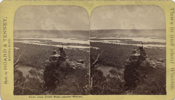 Stereograph of an elevated view of two men posing on Castle Rock. The Mississippi River is below with the city of Winona in the background, and hills are on the horizon. Caption reads: "Views from Castle Rock, opposite Winona." Printed on the reverse: "Views in and about Winona, Minn. Photographed and Published by Hoard & Tenney, No. 18 Center Street." More stereographs in this series are listed.