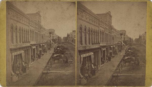 Elevated view of an unpaved main street with storefronts and horse-drawn vehicles hitched along the boardwalk. Many stores have signs and awnings, including "Pettibone's Dry Goods, Carpets & C.," "M. Hawley," "Dentist" and "Doctor Hendricks." A large clock with eyeglasses attached is hanging from a pole in front of "M. Hawley."