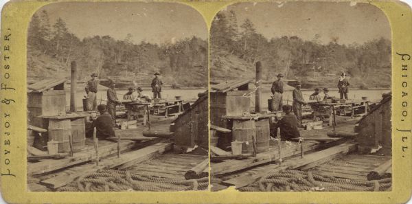 Stereograph of six men on a raft on the Wisconsin River. Structures, barrels, tables on sawhorses, ropes and timbers cover the raft. In the background is the far shoreline, covered with rock formations and trees. Hand-written on the reverse: "Rafting on the 'Wisconsin' River."
