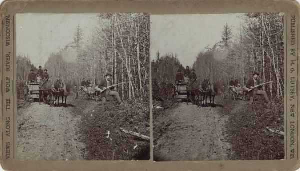 Stereograph of two Menomonee Indian family groups in horse-drawn wagons. One boy is standing next to a wagon and a man is standing on the right holding a gun. A forest surrounds the clearing and dirt road. Printed on the stereograph: "Views along the Wolf River, Wisconsin. Published by H.G. Lutsey, New London, Wis." Text on the reverse indicates that this stereograph is part of the "Menomonie Indian Reservation Series" and the view named "Holzhay's Station."