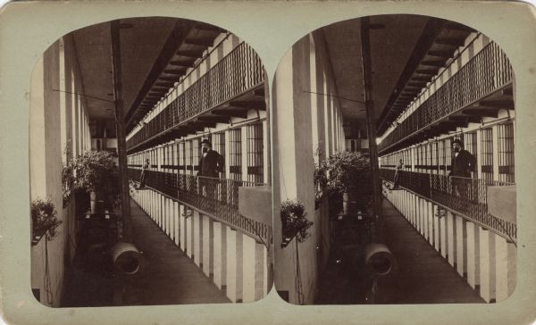 Stereograph of three men posing in a long prison hallway. Two men are on the first level balcony with one on the inside of the railing and the other man standing on a support jutting outwards. The third man is leaning over the second level railing. Cell doors on the first level are open. Square stone columns support the structure. On the left are windows with plants and ductwork. Hand-written on the reverse: "Wisconsin, probably Waupun Wis. By J.C. Sunderland. Cell Rooms (East Side) WI State Prison."