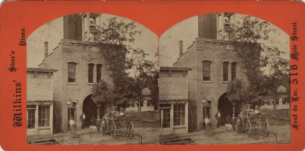 Stereograph of a fire station with a belfry at 516 Main Street. The "No. 1 Steamer" fire engine is parked on the boardwalk. Seven men and two dogs are  standing or sitting on the left of the fire engine. A home is on the right and a store is on the left. Text on front reads: "Wilkins' Stere'o Views. Fond du Lac, 516 Main Street." Text on reverse: "Wilkin's Stereoscopic Views. No. No. 1 Steamer, Published by O.D. Wilkins, Fond du Lac, Wis. Over Hoyt's Shoe Store."