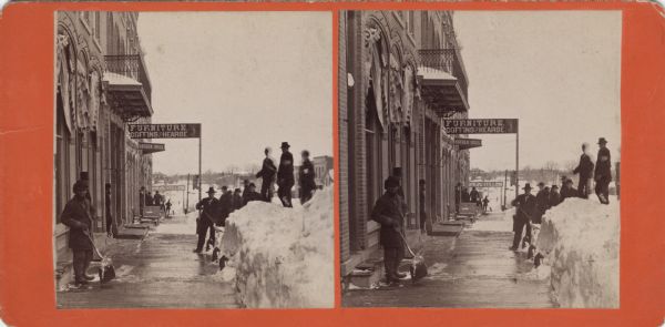 Stereograph of men standing on a cleared sidewalk in front of storefronts. Three men are standing on snowbanks along the curb. Several men are holding snow shovels. Signs on storefronts read: "Furniture. Coffins and Hearse," "Hayden Bros.," "F. Miller City Bakery" and "Sash Doors Blinds Moulding." Text on reverse: Street Scenes After the Great Snow Storm of March, 1881. Columbus and Vicinity. Photographed by W.H. Hosken, Columus, Wis."