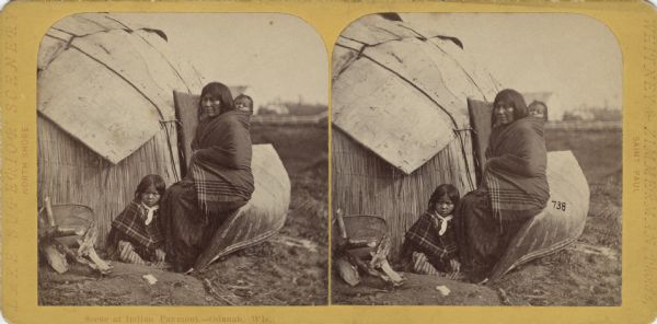 Stereograph of an Ojibwa woman sitting on an overturned canoe with a baby on her back wrapped in a blanket. A young child is sitting at her feet. They are posing in front of a wigwam. In the distance is a farm inside a fence. The caption reads: "Scene at Indian Payment—Odanah, Wis." Text printed on the front: "Lake Superior Scenery. North Shore. Whitney and Zimmerman, Photos, Saint Paul." Hand-written on reverse: "Charles A. Zimmerman. 1866. Bad River Reservation. 410."