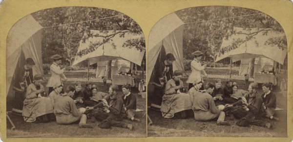 Stereograph of a group of young people gathered in front of a tent. They are engaged in card playing, reading and needlework. In the background is a table under an awning, another tent and many trees. Printed on reverse: "Wm. A. Fermann, Photographer. Stoughton, Wisconsin."