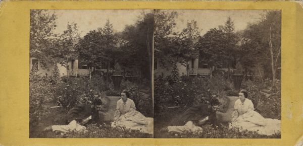 Stereograph of a man and woman relaxing, reclining in the garden of a house. The man is ducking his head under the foliage of a plant behind him. A vase-shaped planter is displayed in the center of a circular garden path. The house is screened by trees with an extensive deck or balcony. Handwritten on reverse: "'Paradise Regained.' Dudley." Printed on reverse: "Manufactured by J.W. Love, Portage City, Wisconsin."