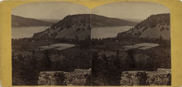 Stereograph of an elevated view of Devil's Lake and the valley to the southeast. The valley may be the site of the old campground that has been moved farther away from the lake. In the foreground is the top of a rock formation.