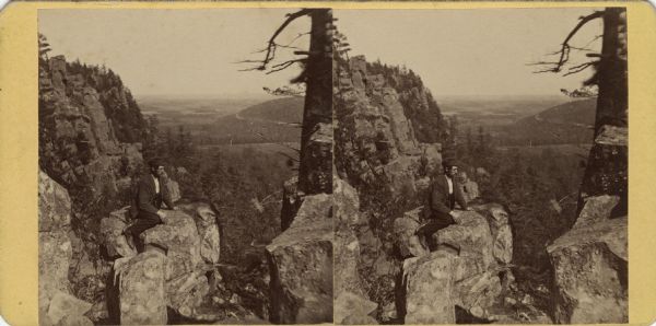Stereograph of an elevated view of a man posing on a rocky outcropping near bluffs, with a valley in the distance. Text on the reverse: "K?iks Valley, Devil's Lake, No. 18., Baraboo, Wis. Photographed by M. Mould."