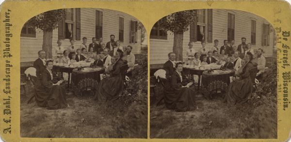 Stereograph of Reverend I.A. Ottesen with his family and visitors sharing outdoor refreshments. On the front table are two stereograph viewers and stereograph cards. Coffee cups are stacked on another table. In the background is a clapboard house with windows and shutters. Text on reverse: "Rev. I.A. Ottesen, Family & Visitors. Utica, Dand Ce., Wis. Andrew L. Dahl, landscape Photographer, DeForest, Wisconsin. Note viewers and cards on table."