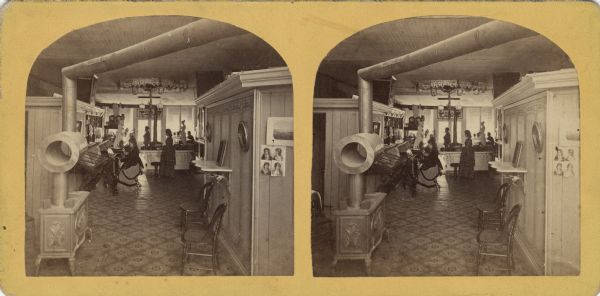 Stereograph of the interior of a jewelry and art store with employees and customers.  This view is from the front of the store towards the rear. In the lower left is a woodstove.<p>Text printed on the back: "Interior View of C.B. Manville's Jew'lry Store and Art Gallery. Established 1861. I have now one of the most complete Establishments of the kind in the Northwest. My stock of Watches and Clocks, Jewelry, Plated Ware, Frames, Chromos and Albums, Is the largest and best in this section. I also sell the <i>McPhail, Jewett & Co. and Root & Cady</i> Pianos. My Prices defy Competition for the same quality of goods. My Photographic Department Is complete, and we are turning out the best o work. Everything new and tasty is to be found here. Thanking you for past favors. I earnestly solicit a share of your patronage, and shall always study to please, and use my best endeavors to make it to your advantage and pleasure to trade with me. Yours, very truly, C.B. Manville. Neenah, Wisconsin."</p>