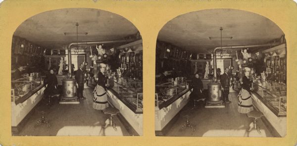 Stereograph of the interior of a jewelry and art store with employees and customers. This view is from the rear of the store towards the front windows. A large woodstove is in the center with glass display cases on both sides.<p>Text printed on the back: "Interior View of C.B. Manville's Jew'lry Store and Art Gallery. Established 1861. I have now one of the most complete Establishments of the kind in the Northwest. My stock of Watches and Clocks, Jewelry, Plated Ware, Frames, Chromos and Albums, Is the largest and best in this section. I also sell the <i>McPhail, Jewett & Co. and Root & Cady</i> Pianos. My Prices defy Competition for the same quality of goods. My Photographic Department Is complete, and we are turning out the best o work. Everything new and tasty is to be found here. Thanking you for past favors. I earnestly solicit a share of your patronage, and shall always study to please, and use my best endeavors to make it to your advantage and pleasure to trade with me. Yours, very truly, C.B. Manville. Neenah, Wisconsin."</p>