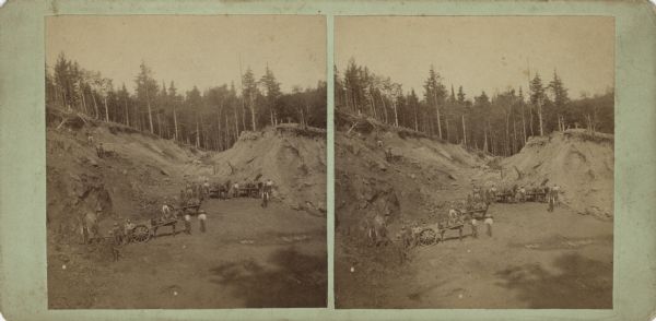 Stereograph of the side view of a mine. Several horse-drawn wagons are waiting for loads with workmen. Hills of soil surround a flat area, and on the horizon are undisturbed trees. The mine entrance is probably on the left. Hand-written on the reverse: "Side view of mine, Florence, Wis."
