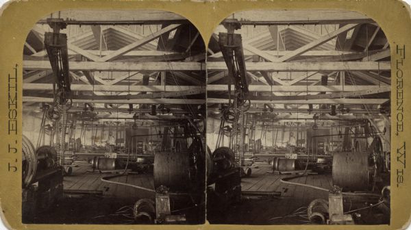 Stereograph of the interior view of a factory. Machinery, materials, gantries, chain hoists, chains, cables, line shafts, and equipment fill the structure. The ceiling is constructed of open trusses.<p>Text on front reads: "J.J. Eskil, Florence, Wis." Text on reverse reads: "A Word Picture of Florence. Florence, county seat of the flourishing new county of Florence, situated in the northeastern portion of Wisconsin, on the line of the Chicago & Northwestern railroad, about 300 miles north of Chicago. Surrounded by the most beautiful works of the great Architect, this new town that has sprung from an unbroken wilderness to a bustling place of 3,000 inhabitants in three years, presents many unusual advantages for obtaining the necessaries [sic] of life, obtaining the wealth of a Croesus or securing and cherishing that greatest of all boons; Perfect health. The air is bracing indeed and healthful in the extreme as it comes wafted through the whispering pines and invigorating balsams, from the bosoms of two great inland seas; Superior and Michigan. The climate is unusually healthy and the evenness of temperature found here is a great rarity the world over. The summers are always cool and the winters dry and unchangeable, Florence county possesses great timber, mineral and agricultural wealth. The season of 1885 will see it traversed by two new lines of railway, the Milwaukee & Northern and Minneapolis, Sault Ste Marie & Atlantic — giving us a competing line to Chicago and an outlet to a vast new territory whose demands are for those articles which we so readily produce and whose excesses are our wants. Iron exists in unlimited quantities and two great mines, which have already shipped hundreds of thousands of tons of ore, have been developed within the county's confines, while many other valuable deposits only await the magic touch of the pick and shovel. Gold, silver, copper and mica have been found in quantities sufficient to lead experienced men to believe that they exist in large deposits in Florence county. The excellent water powers, many useful woods and valuable minerals combine to present a galaxy of splendid manufacturing advantages. Pine abounds, and the valuable hard woods, such as bird's eye maple, go far towards making up our forests. The farmer can obtain good lands at very low prices, the clearing of which will pay for them. He can raise anything congenial to this latitude, such as winter wheat, cats [sic], barley, rye, timothy, clover, potatoes, rutabagos [sic] and in fact all kinds of vegetables, for which he can obtain good market value at home. To the sportsman, tourist or artist this region presents unrivaled attraction. Deer, bear, wolves, lynx, trout, bass pickerel, muscalonge [sic], pheasants, ducks, and many other species of game are here in plenty for the sportsman to dally with. Beautiful lakes, grand forests, picturesque rivers, inspiring glens, romantic canons, majestic dalles, roaring falls and everything else that is interesting in nature exists here, to please the tourist and throw the artist into ecstasy. Then why should people travel thousands of miles from the centres of civilization to obtain less, when all of this honestly exists 300 miles north of Chicago. Florence county possesses better prospects and presents more advantages to settlers than any prairie country and we invite a comparison of the facilities afforded by both. Florence Mining News Print."</p>