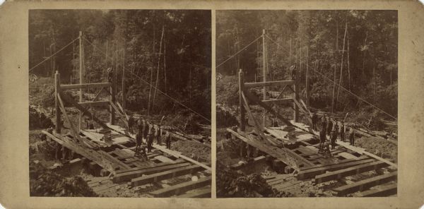 Stereograph of an elevated view of a mining operation. Men are posing on the head frame/gallows frame over the pit. One man is perching on the top of the gallows. There are pulleys, hoists, cables and ropes on the frame. In the background are trees and piles of cut logs. Hand-written on the reverse: "First pocket raised in the Florence mine."