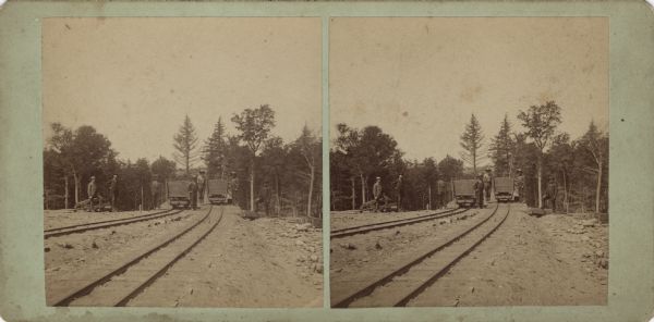 Stereograph of four men standing with ore carts. Two more men are standing on the left near a pile of square timbers, and another man is standing on the right with one foot on a very large timber. The carts are on two sets of railroad tracks crossing a bridge. Trees are in the background.