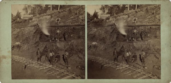 Stereograph of a railroad crew laying tracks. The ties are in place and the rails are being positioned on the first level. The bank behind the crew is terraced, and logs are resting on the third level. The men are standing or sitting on levels one, two and three. On the second level, steam is blowing from a pipe, with more pipe stacked all around. Some buildings can be seen above the fourth level. Hand-written on reverse: "Front view of Florence mine."
