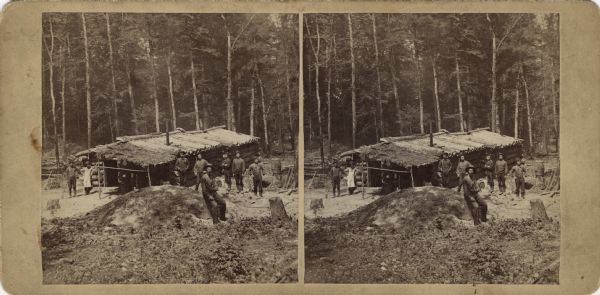 Stereograph of a group of men posing near a log cabin in the woods. In the foreground is a dirt mound and a man sitting on a box. Barrels and tools are scattered about. One man (the cook?) is wearing an apron. Trees are in the background. Text on reverse: "Stereoscopic Views, -of- Florence and Commonwealth. -Published by- Wm. Collins. Neenah, - - Wisconsin."