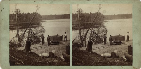 Stereograph of men with an ore cart on railroad tracks. Two men are holding shovels. In the foreground in an earthen bank and tree stumps. There is a lake or river behind them, and a wooded shoreline in the distance. Text on reverse: "Stereoscopic Views, -of- Florence and Commonwealth. -Published by- Wm. Collins. Neenah, - - Wisconsin."