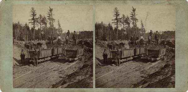 Stereograph of men posing with ore carts on railroad tracks. One man is standing on the dirt in the cart to the right. They are possibly excavating more dirt to extend the track farther. There are earthen banks on both sides. The horizon is filled with trees. Text on reverse: "Stereoscopic Views, -of- Florence and Commonwealth. -Published by- Wm. Collins. Neenah, - - Wisconsin." Hand-written on reverse: "Depot at Florence 1880."