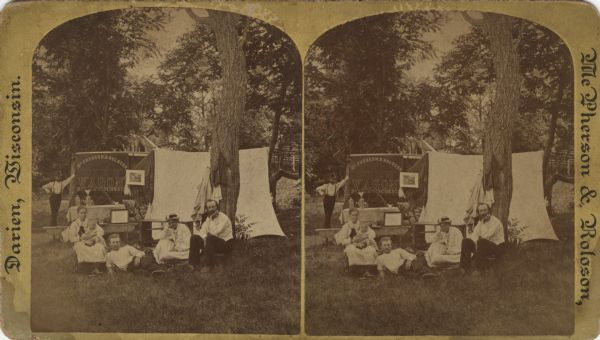 Stereograph of a group portrait of five people, including three men, two women, and an infant, posing on the grass in a wooded area. In the background, on the left, is a "McPherson & Roloson View Wagon, Darien, Wisconsin." On the right is a tent or backdrop. Clothing and blankets are hanging from the poles and trees.