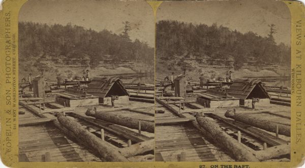 Stereograph of men on a large raft on the Wisconsin River, with exposed, tree-lined rock formations on the shoreline in the background. Wanigans (shelters) and furniture for daily living are set on the raft. Tableware, crocks and basins are sitting on the table. Seven men can be seen near the table. Logs and timbers are pegged to the deck. Text on the card: "Copelin & Son, Photographers, 244 West Washington Street, Chicago. 27. On the Raft. Views at Kilbourn Dalles, Wisconsin."