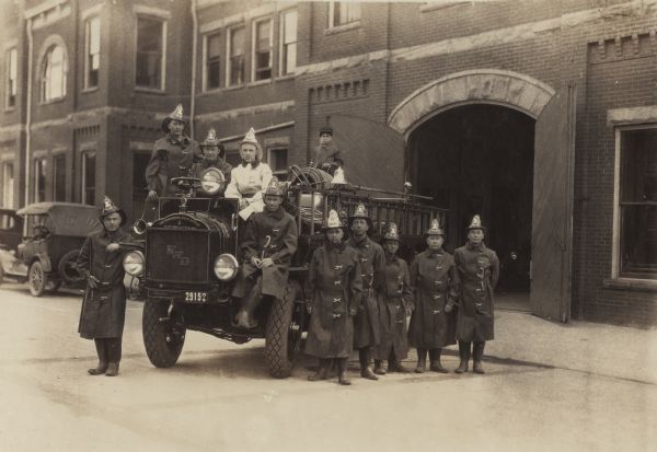 Group portrait of the New London Fire Department posing around their fire engine. They are wearing firefighting gear, coats, boots, helmets and tools in their belts. A boy is standing on the top of the engine behind the fire hose reel next to a bell. The shields on the helmets read: "New London Fire Dept." and the number "1" or "Fire Chief" and "NLFD." The fire engine was made in Clintonville, Wisconsin, by The Four Wheel Drive Auto Company. The plate on the front of the firetruck reads: "The Four Wheel Drive Auto Co., 4, Clintonville, Wis. USA." In the center of the radiator are the capital letters "FWD." The license plate is 2915 WIS.