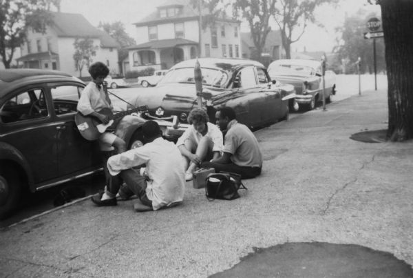 A group of friends relaxing together on Williamson Street. A woman is leaning against a Volkswagon Beetle automobile with a guitar strapped over her shoulder. On the far right sitting on the sidewalk is Lewis Arms, and next to him is Janet Sauk (Hanson).
