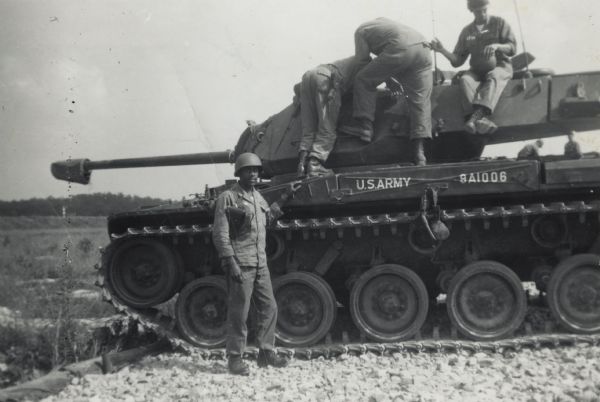 Lewis Arms is standing next to an army tank in Fort Knox, Kentucky. He is wearing a military uniform and helmet. Three men are standing and sitting on top of the tank.