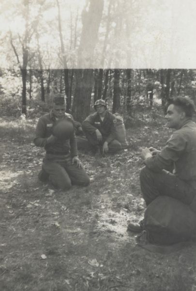 Three soldiers kneeling or sitting on the ground under trees. The soldier on the left is holding his helmet near his chest and is looking downward.