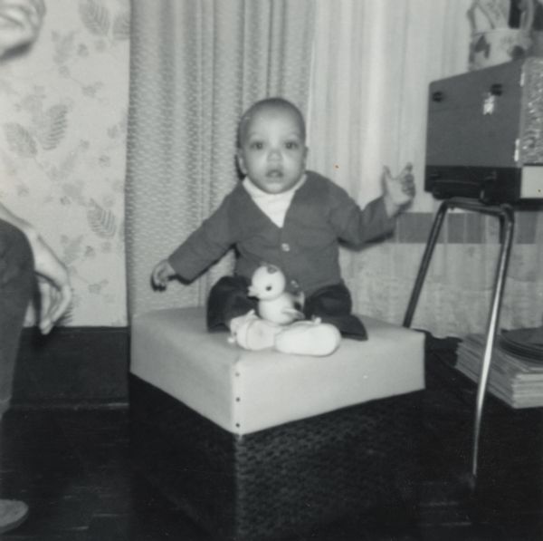 Paul Arms as an infant sitting on an ottoman with a toy on his lap at Lewis Arms' house at 847 Williamson Street. An adult is partially visible on the left.
