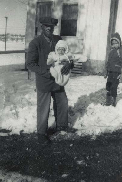 Bernard Arms standing outdoors holding Paul Arms, the baby son of his nephew Lewis Arms. Lewis Arms' daughter Rita is standing behind them. Snow is on the ground.