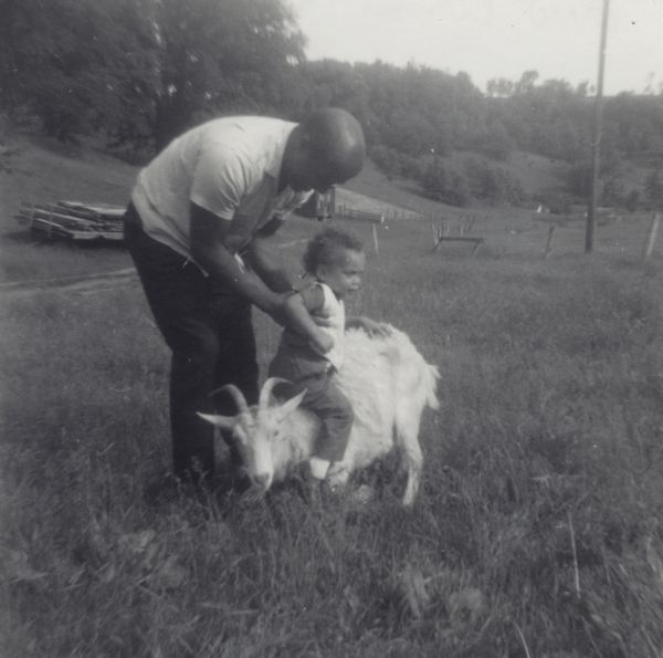 Lewis Arms is holding his toddler son Paul on the back of a billy goat in a grassy field.