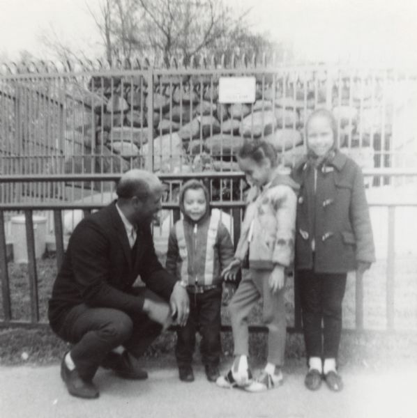 The Arms family posing together in front of an animal enclosure at Vilas Park Zoo. From left to right are Lewis Arms, and his children Paul Arms, Mamie Arms, and Rita Arms.