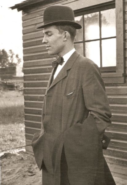 Three-quarter length portrait of Harry Lynde Bradley as a young man, standing posing outdoors in front of a building. He is wearing a suit and hat, and has a coat over one arm.