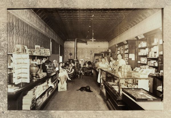 Interior view of the drugstore owned by Jerome Francis Franklin Jr., located next to the Franklin Hardware Company/Eland Post Office. In the foreground are counters on both sides displaying merchandise. On the right is Elsie Franklin, the wife of the owner, and two of their daughters standing at the counter. The woman on the far left is another daughter. Their names are Belle, Nola and Joyce. In the middle, a dog is lying on the floor near a sign advertising Carver ice cream. In the background is a seating area with tables, filled with customers, and a woodstove.
