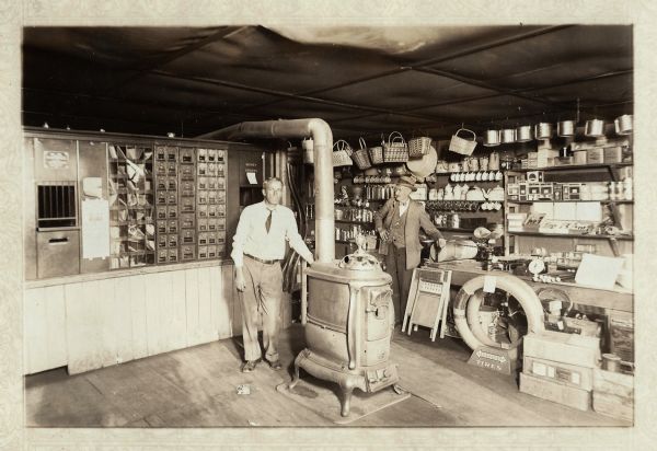 Two men posing in the interior of the Franklin Hardware Company and Eland Post Office. Jerome Francis Franklin Jr. is on the left, Jerome Francis Franklin Sr. is on the right. Between them is a coal burning parlor stove. Merchandise fills the floor and shelves on the right. On the back wall, post boxes can be seen. Jerome Francis Franklin Sr. and his son Jerome Francis Franklin Jr. alternated in the job of Postmaster.