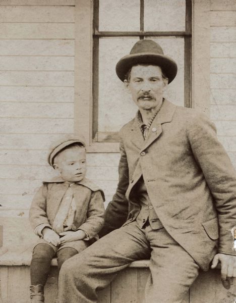 Informal outdoor portrait of Jerome Francis Franklin, Sr. and his son Perry Small Franklin. They are seated on a box in front of a wall with a window.