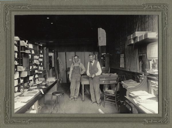 An unknown man and Jerome Francis Franklin, Sr. (right) pose in the back room office of the Franklin Hardware Company and Eland Post Office. On the left are the open backs of the post boxes and a long counter with a stool. On the left are shelves, a table, chair and desk. Jerome Francis Franklin Sr. and his son Jerome Francis Franklin Jr. alternated in the job of Postmaster.