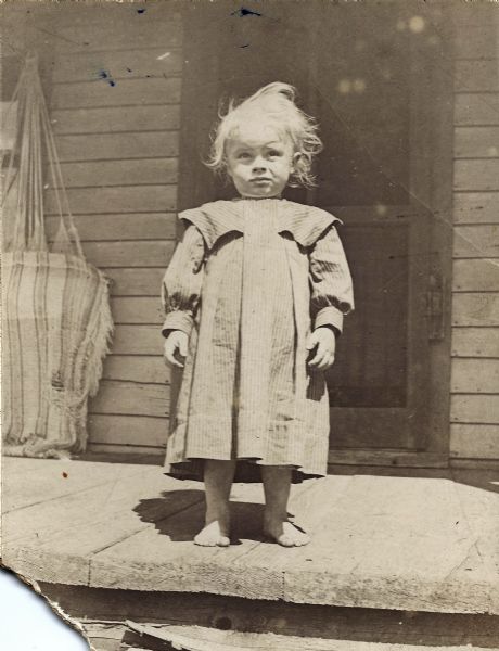 Perry Small Franklin standing on the porch of a house, a door in the background. He is approximately two years old. He is wearing a short dress, the kind young children of both sexes wore for their everyday clothes until about age 2-4. A tattered hammock hangs on the wall on the left.