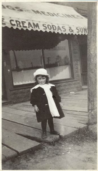 Alta Belle Franklin standing on the boardwalk outside of her father, Jerome Francis Franklin Jr.'s, Drug Store. She is wearing a dress, coat and hat. Text on the awning reads: "Drugs and Medicine" and "Ice Cream Sodas & Sundaes."