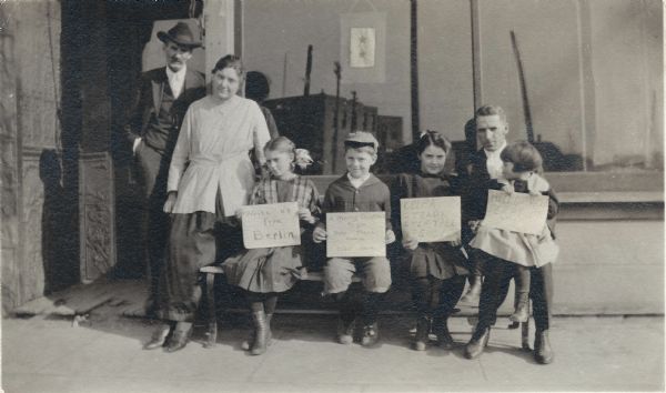 The extended Franklin family posing in front of a store (possibly the family owned Drug Store or Hardware Store) with messages wishing Perry Small Franklin well during his service overseas during WWI. He is the brother of Jerome Francis Franklin Jr. Names (l to r) Jerome Francis Franklin Sr., Elsie Franklin (Jerome Jr's wife), their children, Nola, Hervy, Joyce and Belle on the lap of Jerome Francis Franklin Jr. Two stars are on the banner in the window. One star is for Perry Small Franklin. The other may be for Hervy Edward Franklin, another brother. The signs read: "Write Us From Berlin," "A Merry Christmas To You Over There From Us Over Here," "Keep A Steady Step Till Its Over" and "Hello Uncle Perry."